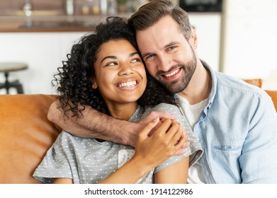 Hilarious multiracial couple in love sits in embraces in cozy living room at home. A close-up portrait of smiling caucasian guy and charming african girl are hugging. Love and affection concept - Shutterstock ID 1914122986