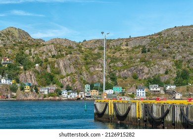 The hillside of St. John's Harbour, Newfoundland, on a sunny day, under blue sky and white clouds. The colorful wooden houses are scattered along the hillside with the blue ocean in the foreground. 