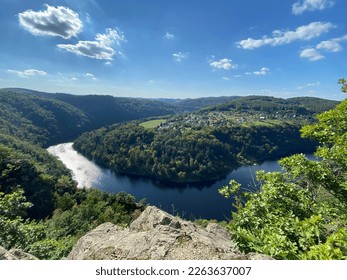 Hillside views over thick woodlands around a dramatic, horseshoe-shaped meander of the Vltava River with a small village located opposite the view during the summer with blue sky. 