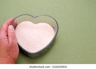 Himalayan salt in a heart shape clear glass dish held by a human adult male hand on a green tablecloth. Heart shaped clear glass dish with Himalayan pink salt held by an adult white male hand.