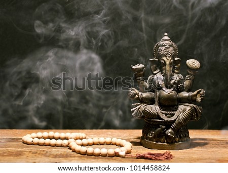 Hindu god Ganesh on black background. Statue and rosary on wooden table with a smoke of incense. Copy space.