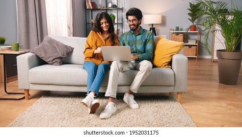 Hindu happy cheerful young married couple sitting on sofa in modern room browsing on laptop computer buying making purchase with credit card, internet shopping, family time, e-commerce concept - Powered by Shutterstock