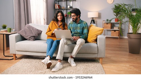 Hindu happy cheerful young married couple sitting on sofa in modern room browsing on laptop computer buying making purchase with credit card, internet shopping, family time, e-commerce concept - Shutterstock ID 1953246964