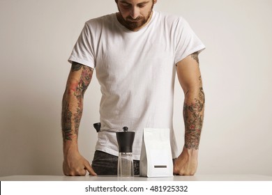 Hip tattooed barista in plain white t-shirt looking down at a small manual burr grinder and unlabeled white bag with coffee beans - Shutterstock ID 481925332