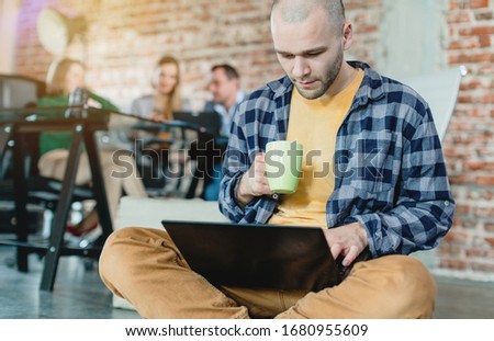 Hip worker in a startup coding with laptop sitting on skateboard with colleagues in background