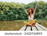 Hippie eco-activist woman traveler sits on a bridge by a lake with her arms outstretched with a hat and smiling sincerely