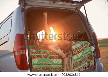 a hippie girl lies upside down and upside down in a minivan in an open field against the backdrop of sunset. High quality photo
