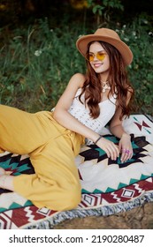 Hippie woman smiling in eco clothing yellow pants, white knit top, hat and yellow glasses sitting on plaid in park watching sunset, lifestyle camping trip