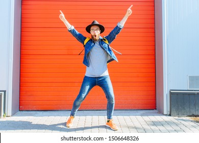 Hipster beard man in eyeglasses, black hat, denim costume, yellow backpack jumping, dancing, having fun, celebrating success with hands in the air and walking on red background. Full height outdoor