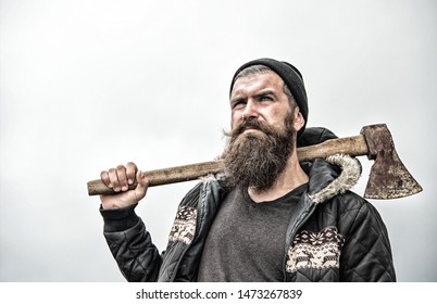 Hipster with beard on serious face carries axe on shoulder sky on background, copy space. Lumberjack brutal and bearded holds axe. Brutal lumberjack concept. Man in hat and warm jacket looks brutally.
