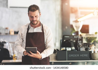 Hipster Bearded Male Barista Holding And Looking At Digital Tablet At The Coffee Shop Or Restaurant Cafe. Startup Of Small Business Owner. Entrepreneur, SME.