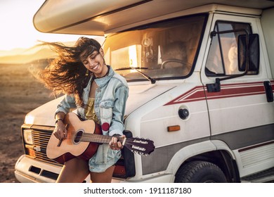 Hipster beautiful woman playing guitar with big smile on her face. Nomadic and camper lifestyle concept. Warm sunset light. 