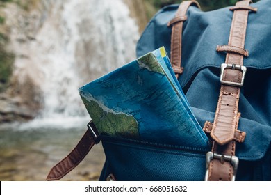 Hipster Blue Backpack, And Map Closeup. View From Front Tourist Traveler Bag On Waterfall Background. Exploring Adventure Hiking Concept