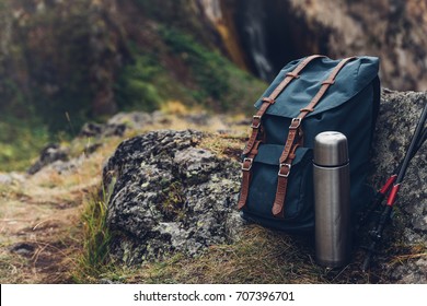 Hipster Blue Backpack, Thermos And Trekking Poles Closeup, Front View. Tourist Traveler Bag On Rocks Background. Adventure Hiking Outdoor Concept