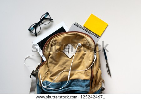 Hipster brown-blue textile backpack, full of school supplies, blank screen tablet, earphones, glasses, notebook, yellow stickers on white background. Back to school concept. Close up, copy space, flat