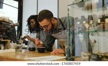 Hipster caucasian barista with beard and african young woman working in modern trendy coffee shop cafe, woman washing cup and man sniffing the fresh coffee.