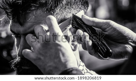 Hipster client getting haircut. Hands of barber with hair clipper, close up. Haircut concept. Man visiting hairstylist in barbershop. Barber works with hair clipper. Black and white.
