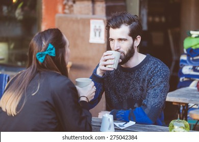 Hipster Couple Drinking Coffee In Stockholm Old Town. They're Sitting Face To Face. The Man Is Wearing A Blue Sweater And The Woman A Striped Shirt With Black Leather Jacket.