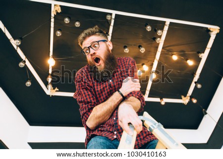 Hipster creative beard guy in eyeglasses balancing on construction worker climbing a ladder while working on home interior over black ceiling background. Anxiety unhappy expression concept.