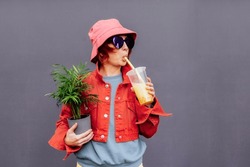 Hipster Fashion Woman In Bright Clothes, Heart Shaped Glasses, Bucket Hat Drinking Fruity Sugar Flavored Tapioca Pearl Bubble Tea And Holding Green Potted Plant On The Gray Wall Background.