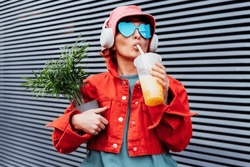 Hipster Fashion Woman In Bright Clothes, Heart Shaped Glasses, Headphones, Bucket Hat Drinking Fruity Flavored Tapioca Bubble Tea And Holding Green Potted Plant On The Gray Striped Wall Background.