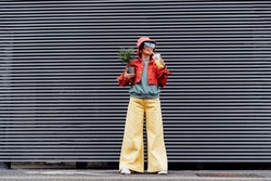 Hipster Fashion Woman In Bright Clothes, Heart Shaped Glasses, Bucket Hat Drinking Fruity Sugar Flavored Tapioca Pearl Bubble Tea And Holding Green Potted Plant On The Gray Striped Wall Background.