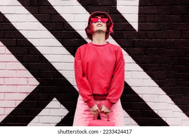 Hipster fashion young woman in trendy magenta color sweatshirt and sunglasses and bucket hat posing on the painted brick wall background. Color of the 2023 year. Urban city street fashion. - Shutterstock ID 2233424417