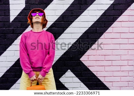 Hipster fashion young woman in trendy viva magenta color sweatshirt and sunglasses and bucket hat posing on the painted brick wall background. Color of the 2023 year. Urban city street fashion.