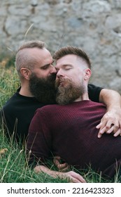 Hipster Fashionable  Gay Couple Kissing In Grass.  Long Beard Male Homosexual Partners Lying Together Outside, Holding Hands, Enjoying In Romance Outdoors. 