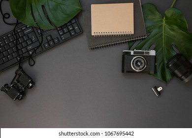 Hipster Flatlay With Camera, Keyboard, Lens, Monstera Leaves, Notebook And Binocular On Grey Table. Dark Working Space Top View. Creative Work Table For Photographer, Traveller, Blogger