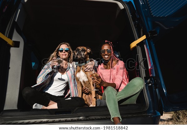 Hipster friends with dog celebrating with wine\
together in camper van during summer travel vacation - Main focus\
on african girl face