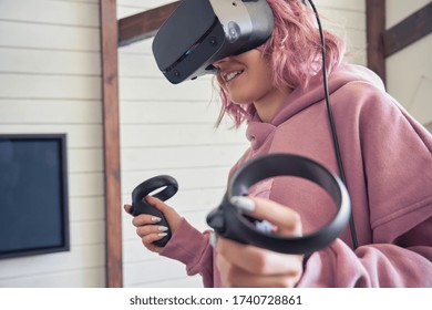 Hipster gamer teen girl pink hair wear vr headset goggles hold handheld controllers play video game simulator immersive futuristic virtual reality 3D vr travel tour, watch 360 video, stand at home.