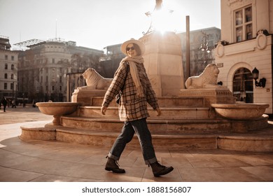 Hipster girl in beige hat, jeans, checkered coat and sunglasses ties scarf and walks around beautiful old European city.