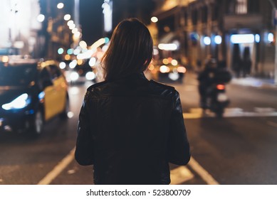 Hipster girl in black leather jacket from back on background illumination glow bokeh light in night atmospheric christmas city, hipster costs at a stop in headlights auto taxi; mockup glitter