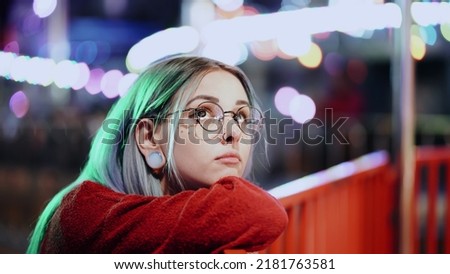 Hipster girl with blue dyed hair, golden sequins as freckles. Sad woman with nose piercing, transparent glasses, ears tunnels, unusual hairstyle stands in amusement night park. High quality photo