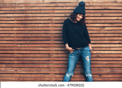 Hipster girl with curly hair wearing black sweater, hat and jeans posing against wooden wall, swag street style, autumn outfit
