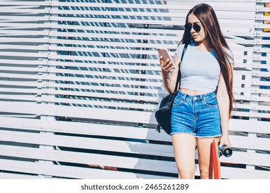 Hipster girl dressed in casual outfit reading incoming notification from network on smartphone holding skateboard in hands while standing outdoors on promotional background for advertising text - Powered by Shutterstock