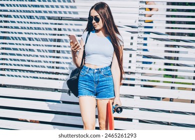 Hipster girl dressed in casual outfit reading incoming notification from network on smartphone holding skateboard in hands while standing outdoors on promotional background for advertising text - Powered by Shutterstock