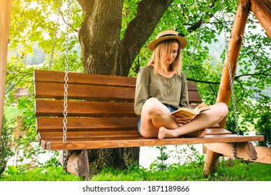 Hipster Girl In Glasses Is Reading A Book On Wooden Bench. Woman Relaxing Outdoors On Porch Swing In The Nature. Cozy Photo. Summer Mood.