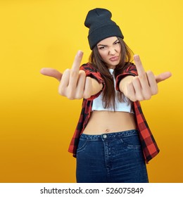 Hipster girl in jeans, checked shirt and hat showing middle fingers over yellow background. Impertinent behaviour. Hipsters. Provocation. Aggression. Naughtiness.
