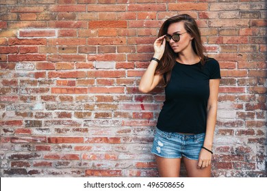 A hipster girl with long brown hair wearing a blank black t-shirt is looking aside while standing on a light blue wooden fence background on a street.Horizontal mock up. Empty space for text o design.