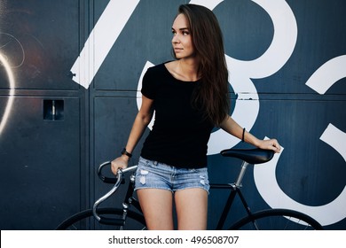 A hipster girl with long brown hair wearing a blank black t-shirt is looking aside while standing on a light blue wooden fence background on a street.Horizontal mock up. Empty space for text o design.