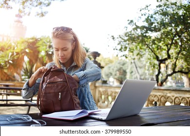 Hipster Girl Looking For Something In The Bag While Sitting Outdoors At The Table With Open Net-book, Young Female Searching The Pen In Rucksack For Writing An Essay On A Notebook From Laptop Computer