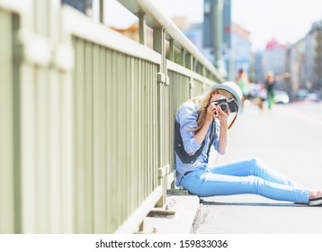 Hipster girl making photo with retro camera while sitting on city street