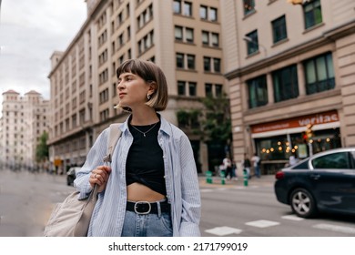 Hipster girl in shirt posing with short hairstyle outdoors. Brunette woman in blue shirt and jeans with brown backpack looking aside in city.