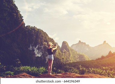 Hipster girl is shooting video of beautiful sundown on cell telephone, while is standing against Amazon scenery background with copy space. Female tourist is taking photo with mobile phone camera