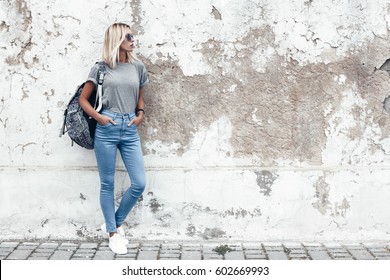 Hipster girl wearing blank gray t-shirt, jeans and backpack posing against rough street wall, minimalist urban clothing style, mockup for tshirt print store