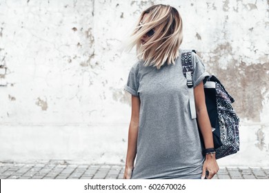 Hipster girl wearing blank gray t-shirt and backpack posing against rough street wall, minimalist urban clothing style, mock up for tshirt print store