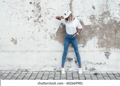 Hipster girl wearing blank white t-shirt, jeans and baseball cap posing against rough street wall, full length portrait, minimalist urban clothing style, mockup for tshirt print store