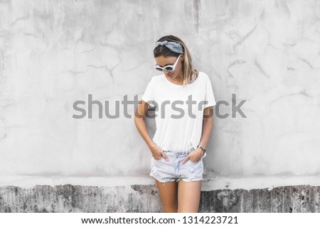 Hipster girl wearing blank white t-shirt and denim shorts posing against gray street wall, blank mockup for tshirt print store